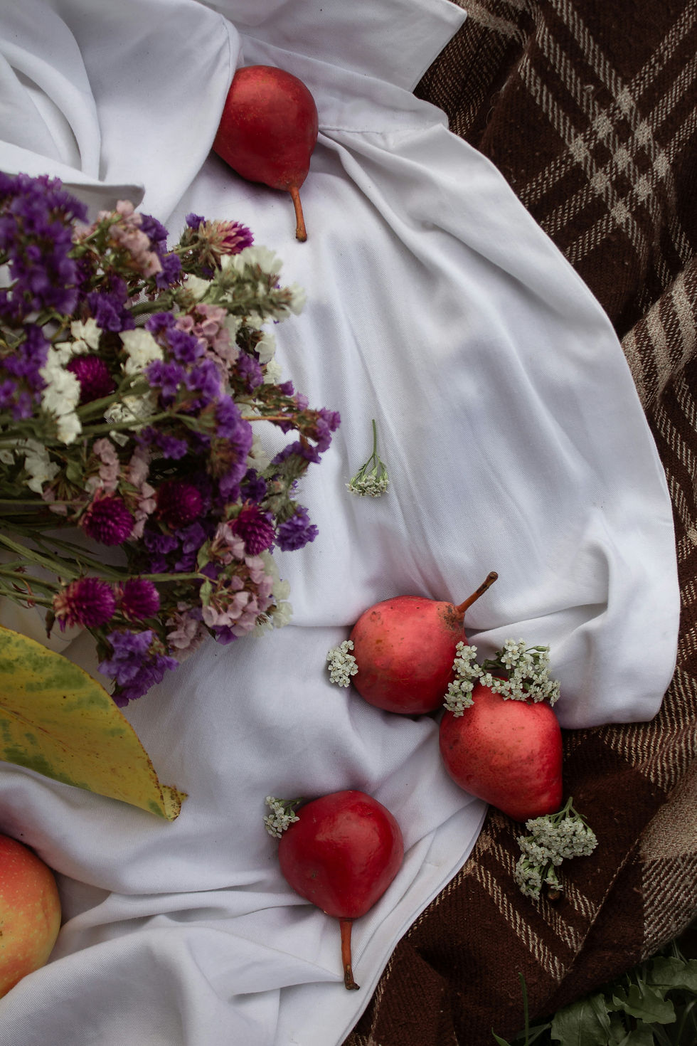 Red pears and purple flowers on white fabric over brown plaid. A yellow leaf adds contrast, creating a rustic autumn scene.