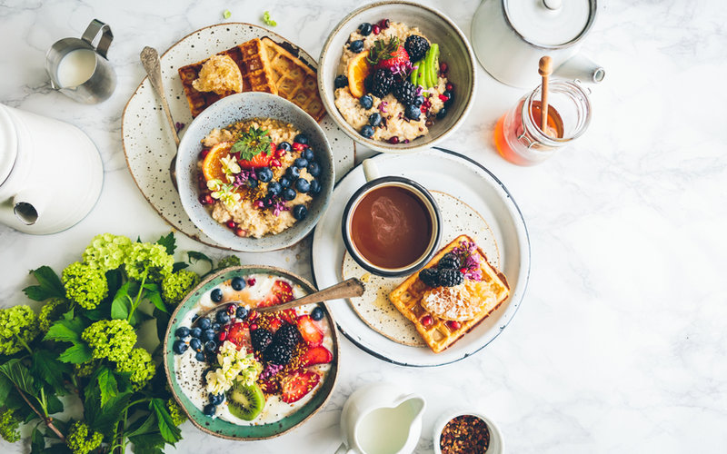 Overhead flat-lay of a colorful brunch spread on a white marble surface with bowls of fruit, waffles, berries, yogurt, coffee, and fresh greens