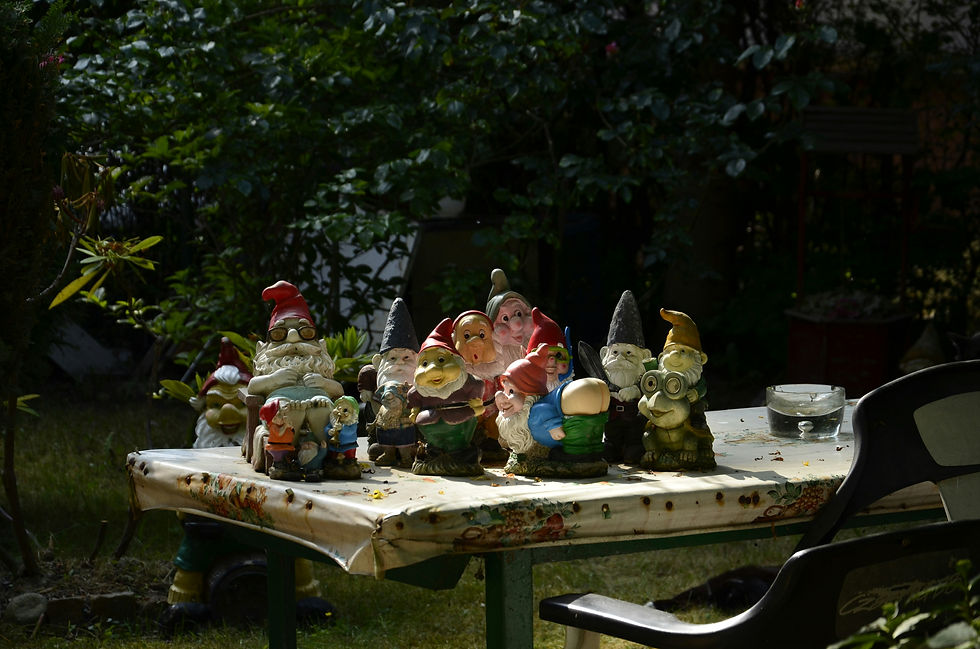 Garden gnomes with colorful hats are displayed on a decorated table in a lush garden. Sunlight casts shadows, creating a whimsical scene.