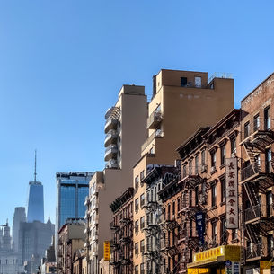 Street view of Lower Manhattan with classic brick buildings and fire escapes in the foreground, Chinatown signage along the block, and One World Trade Center rising in the distance under a clear blue sky.