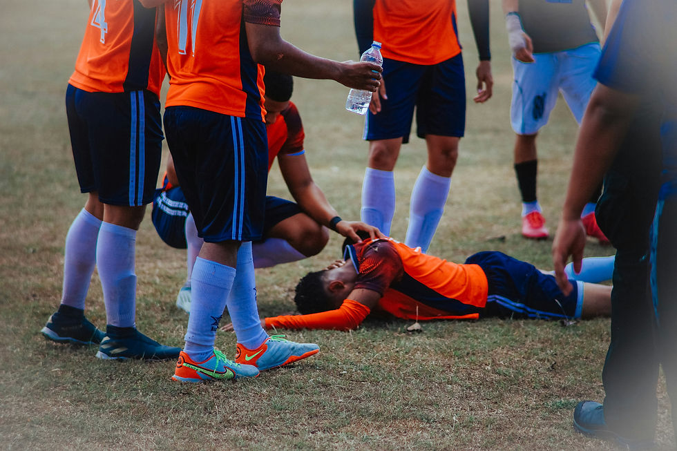 Soccer players in orange jerseys surround an injured teammate on the field, one holding a water bottle. Concerned expressions, grassy background.