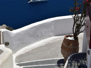 A high-angle view looking down at a white catamaran sailboat with red accents floating on deep blue water, framed by a whitewashed stone wall and a terracotta potted plant with pink flowers in the foreground.
