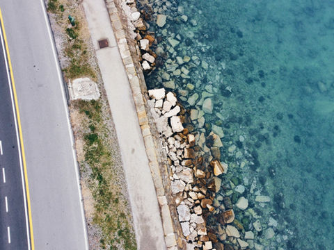Aerial view of a coastal road beside a rocky shoreline and clear turquoise water, with yellow lane lines and no people.