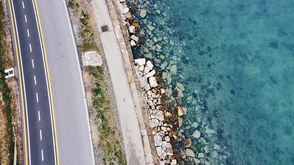 Aerial view of a coastal road beside a rocky shoreline and clear turquoise water, with yellow lane lines and no people.