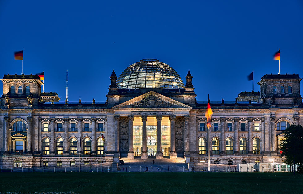 Illuminated building with modern glass dome at night, German flags visible. Text reads "Dem Deutschen Volke." Clear blue sky.