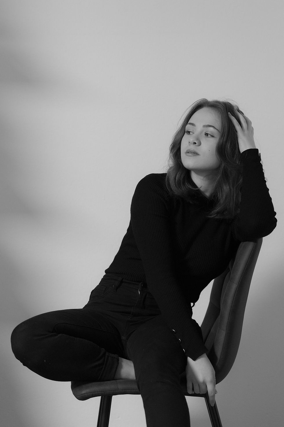 Woman in a black outfit sits on a chair against a plain background, looking contemplative with her hand in her hair. Black and white.