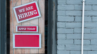 Rote Schilder an Holzwand und grauer Backsteinwand. Text: "WE ARE HIRING" und "APPLY TODAY". Einladende Stimmung.