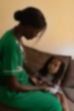 Nurse in green scrubs holds hands with a young girl on a sofa. The girl looks up attentively. Warm, softly lit room. No visible text.