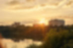 Sunset over a cityscape with buildings and trees, reflecting on a calm lake. The sky glows with warm orange and yellow hues.