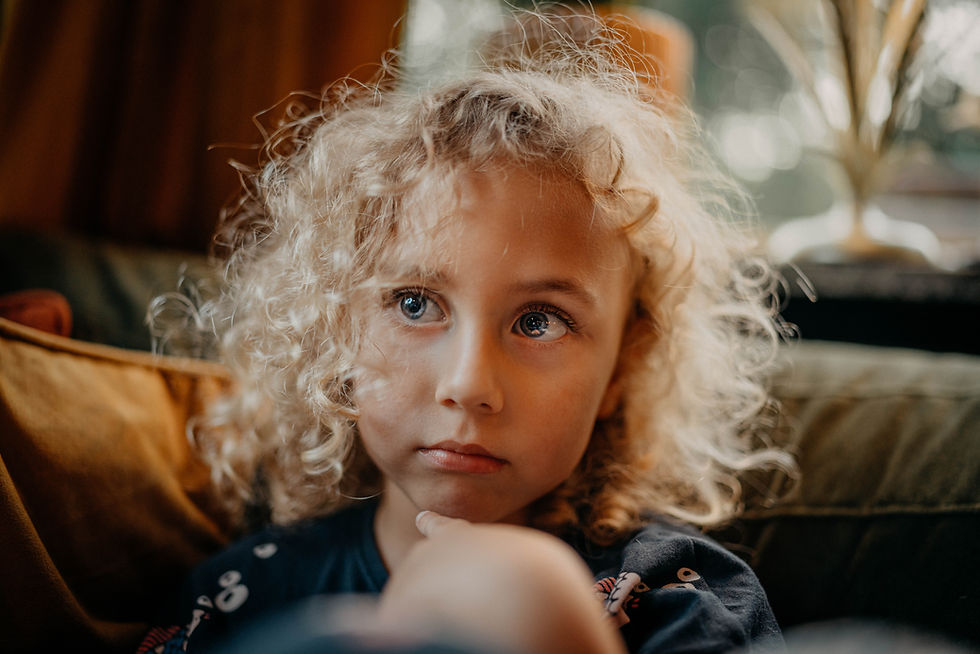 Young child with curly hair gazes thoughtfully off-camera, sitting indoors on a couch. Warm, soft lighting creates a calm atmosphere.