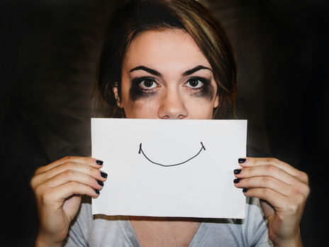 A woman holds a paper smile in front of her face while her eyes reveal exhaustion and emotional strain, symbolizing hidden burnout and the difference between appearing fine and truly thriving.