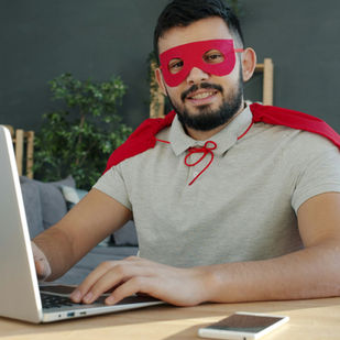 Man wearing a red mask and cape smiles while using a laptop at a desk with a smartphone. Green plants in the background, casual setting.