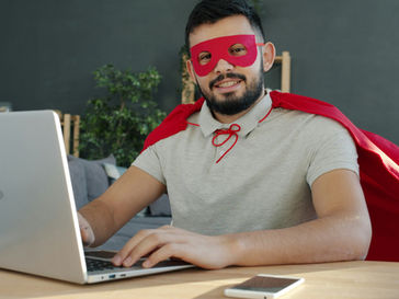 Man wearing a red mask and cape smiles while using a laptop at a desk with a smartphone. Green plants in the background, casual setting.