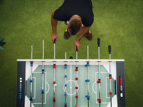 Top-down view of a man playing foosball on green turf, gripping two handles over a red-and-blue table.
