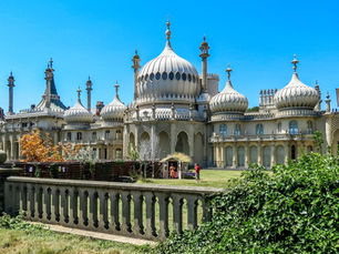 Ornate palace with domes and spires under clear blue sky. Two people in a garden with green shrubs and a stone balustrade in foreground.