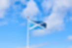 Scottish flag waves on a white pole against a bright blue sky with scattered clouds. The scene is vibrant and peaceful.
