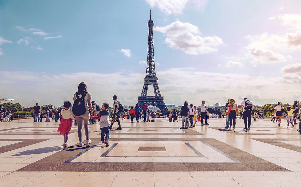Tourists walk across a bright plaza toward the Eiffel Tower under a blue sky with scattered clouds.