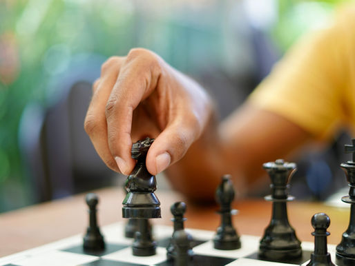 Hand in a yellow shirt moves a black chess knight on a board, with other pieces blurred and a soft green background.