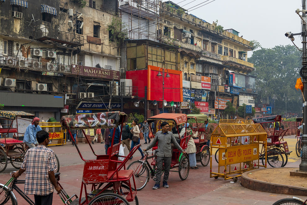 Busy street scene with rickshaws and people in front of old buildings in India. Red and yellow tones dominate, with signs reading Delhi Police.