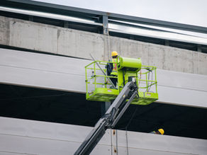trabalhadores realizando um serviço em altura na fachada de um prédio. Um dos profissionais está em uma plataforma elevatória verde (tipo cesto aéreo), suspensa por um braço mecânico articulado. Ele utiliza equipamentos de segurança, como capacete amarelo e colete refletivo, indicando que se trata de uma atividade de construção ou manutenção. Parece estar operando algum equipamento — possivelmente um lavador de alta pressão ou ferramenta para limpeza/manutenção da estrutura externa. Ao lado, há outra plataforma elevatória com mais um trabalhador, também utilizando capacete, sugerindo trabalho em equipe. O prédio tem estrutura de concreto aparente e grandes aberturas horizontais, possivelmente sendo uma obra em andamento ou um edifício comercial/industrial em fase de manutenção. A cena transmite a ideia de trabalho técnico especializado e cuidado com segurança em atividades realizadas em altura.
