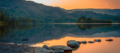 A tranquil lake at sunset, reflecting surrounding hills and trees under an orange sky. Smooth stones lead into the water from a pebbled shore in the foreground.
