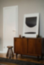 Mid-century style room with a teak cabinet, abstract art leaning against the wall, a small stool, and decor items. Calm, neutral tones.
