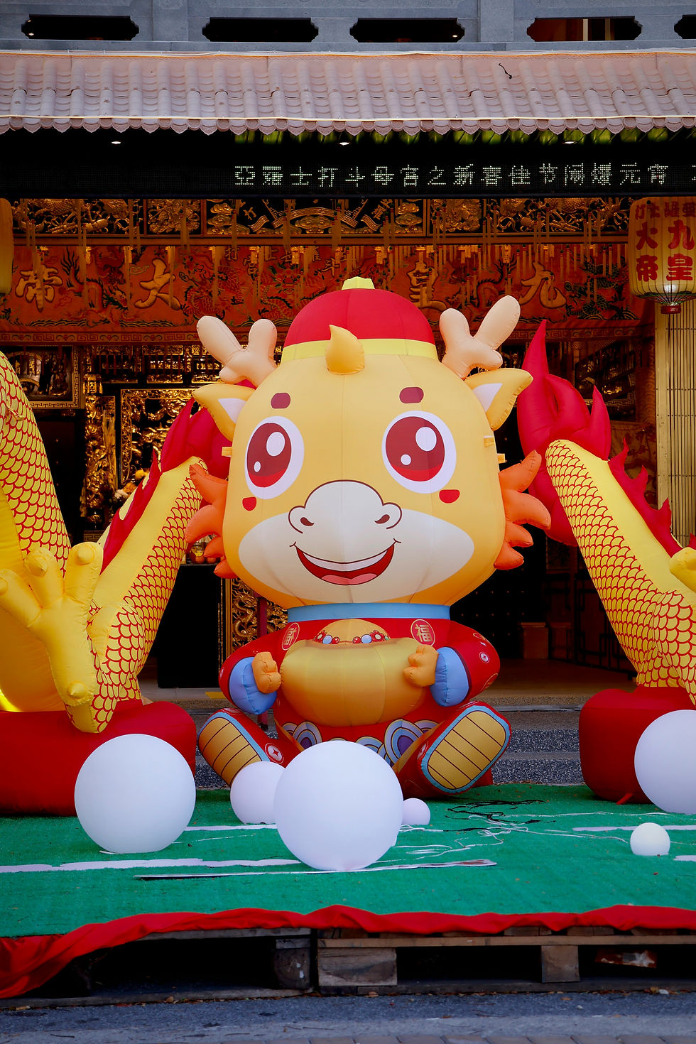Bright inflatable dragon with red and yellow features, smiling on a green platform. Background features gold decor and Chinese text.