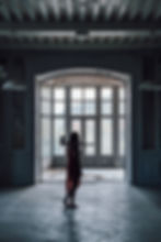 A woman in a red dress stands in an abandoned room, silhouetted against large windows while holding an axe over her shoulder.