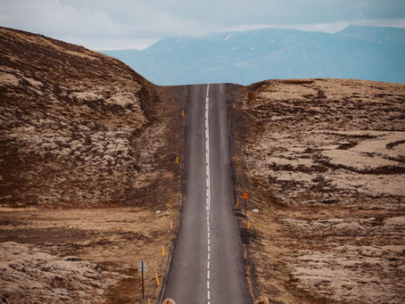 Two people in dark coats walk on a desolate road flanked by rocky terrain, under a cloudy sky with distant mountains.