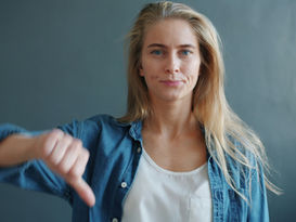 Woman with blond hair in a denim shirt gives a thumbs-down gesture, appearing displeased. Grey background adds a neutral tone.