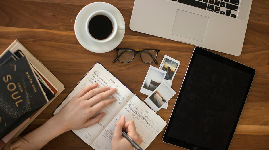 Your Soul book, persons hands writing a journal, coffee cup, glasses, photos, Macbook and tech