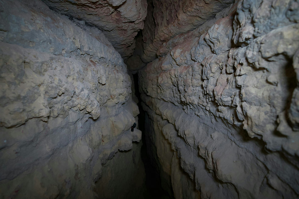 Narrow cave passage with rough, rocky walls in shades of gray and beige. Dimly lit, creating a mysterious, enclosed atmosphere.