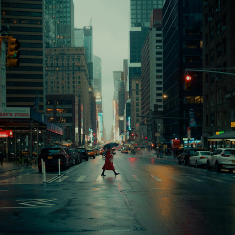 Rainy city street with a woman in a red coat crossing under an umbrella, surrounded by tall buildings, neon signs, and red lights.