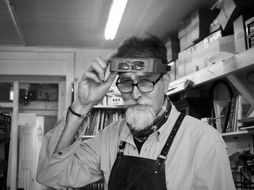 Man in workshop lifts magnifying goggles, wearing glasses and apron. Background has shelves with boxes and books. Monochrome, focused expression.