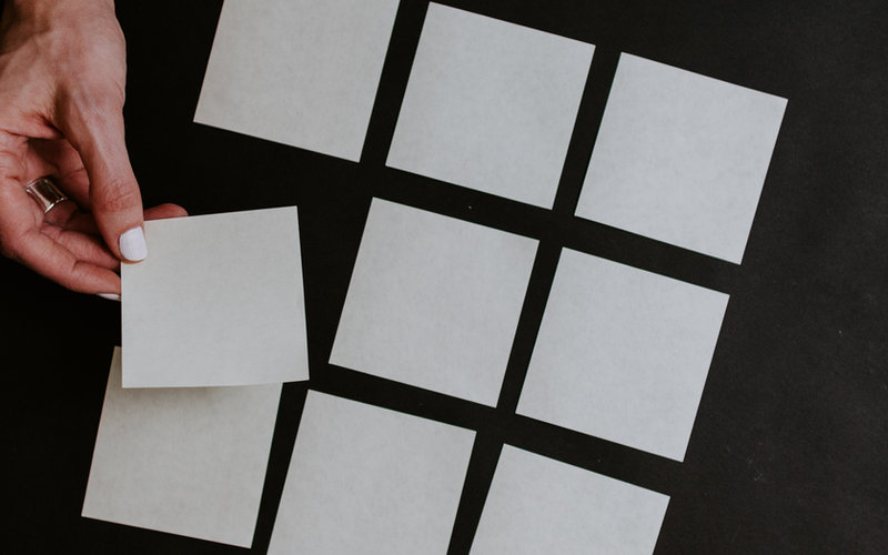 Hand with white-painted nails arranging blank white sticky notes in a precise grid pattern on a black surface