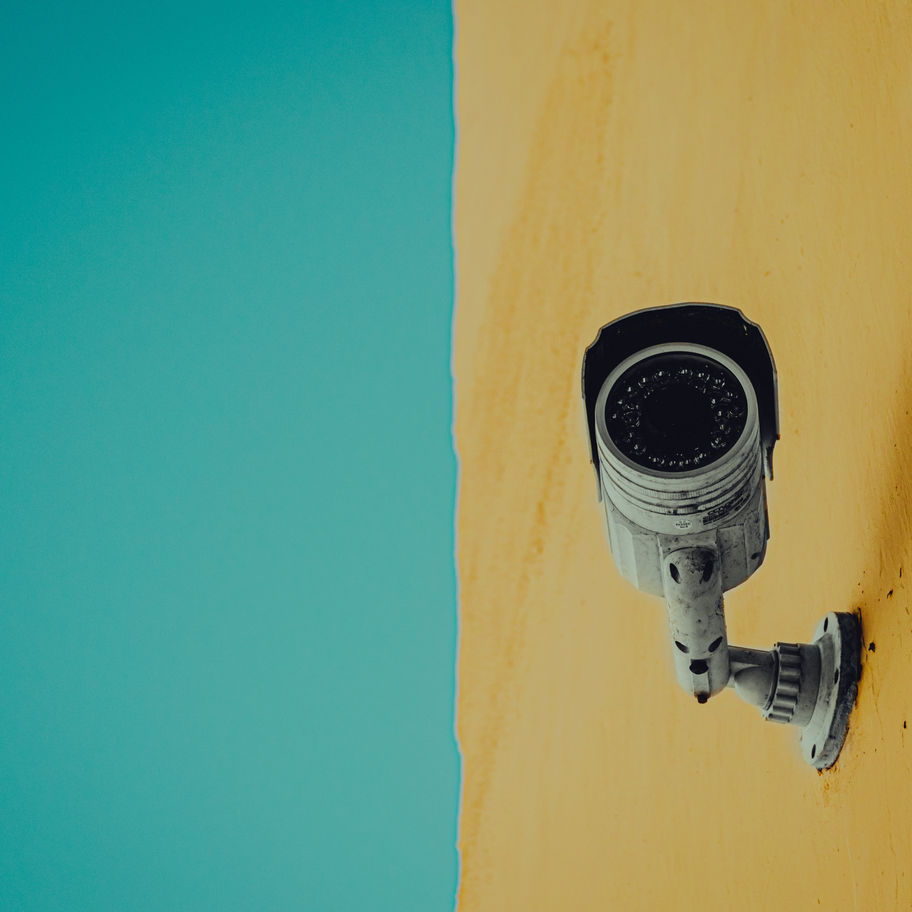 Surveillance camera mounted on a yellow building against a blue sky, symbolizing constant monitoring and essential security.
