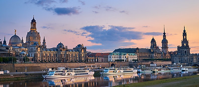 Panoramic view of Dresden's historic skyline at sunset, with ornate buildings and church spires along the Elbe River. Several white riverboats are docked at the riverbank, reflecting in the calm water.