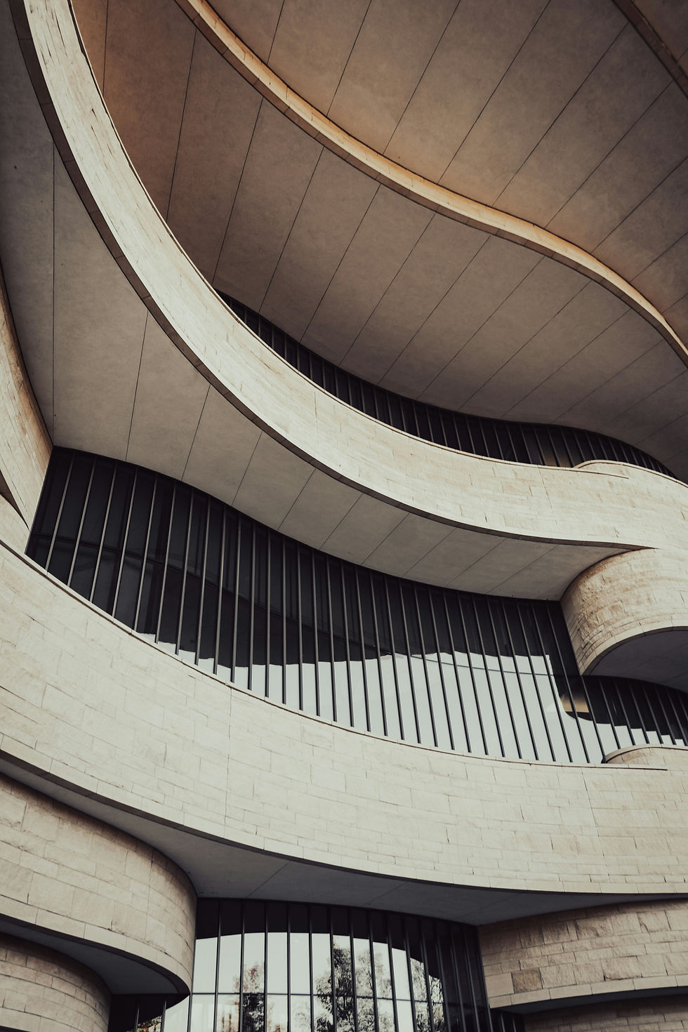 Curved beige stone balconies and dark vertical windows on a modern building facade, seen from below under overcast light.