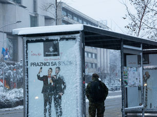 Besneeuwde bushalte met man in camouflagejas; advertentie met Promenada en Praznici, in winterse stadsstraat.