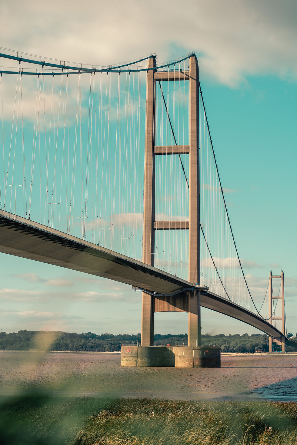 Suspension bridge over river, with blue sky and clouds. Towering structure with cables, surrounded by green landscape and calm water.