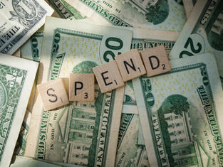 Scrabble tiles spelling "SPEND" on scattered US dollar bills. The setting suggests financial spending, with a focus on money and cost.