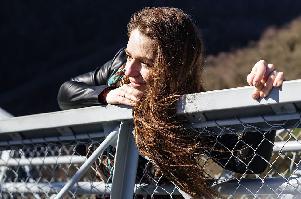 Woman leaning on a metal railing with thoughtful expression. Wind blows her long brown hair. Background features blurred trees.