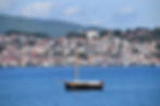 Segelboot auf blauem Wasser vor einer Stadt mit roten Dächern, umgeben von grünen Hügeln und blauem Himmel. Ruhige, friedliche Atmosphäre.