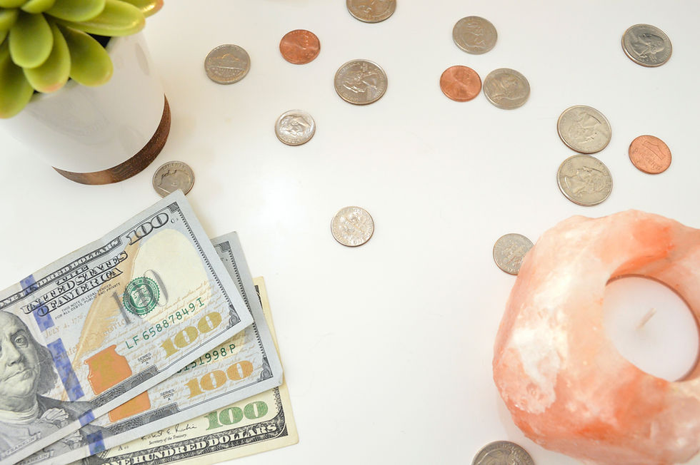 Coins and hundred-dollar bills on a white surface, with a green potted plant and pink salt candle holder nearby.