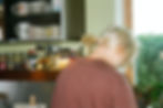 A person with blonde hair faces away in a kitchen. Shelves with spices and mugs are visible. Sunlight streams through a window nearby.
