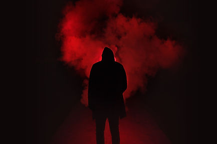 Silhouette of a hooded figure standing in front of a cloud of red smoke, surrounded by darkness—conveying mystery, danger, and the atmospheric tension of horror storytelling.