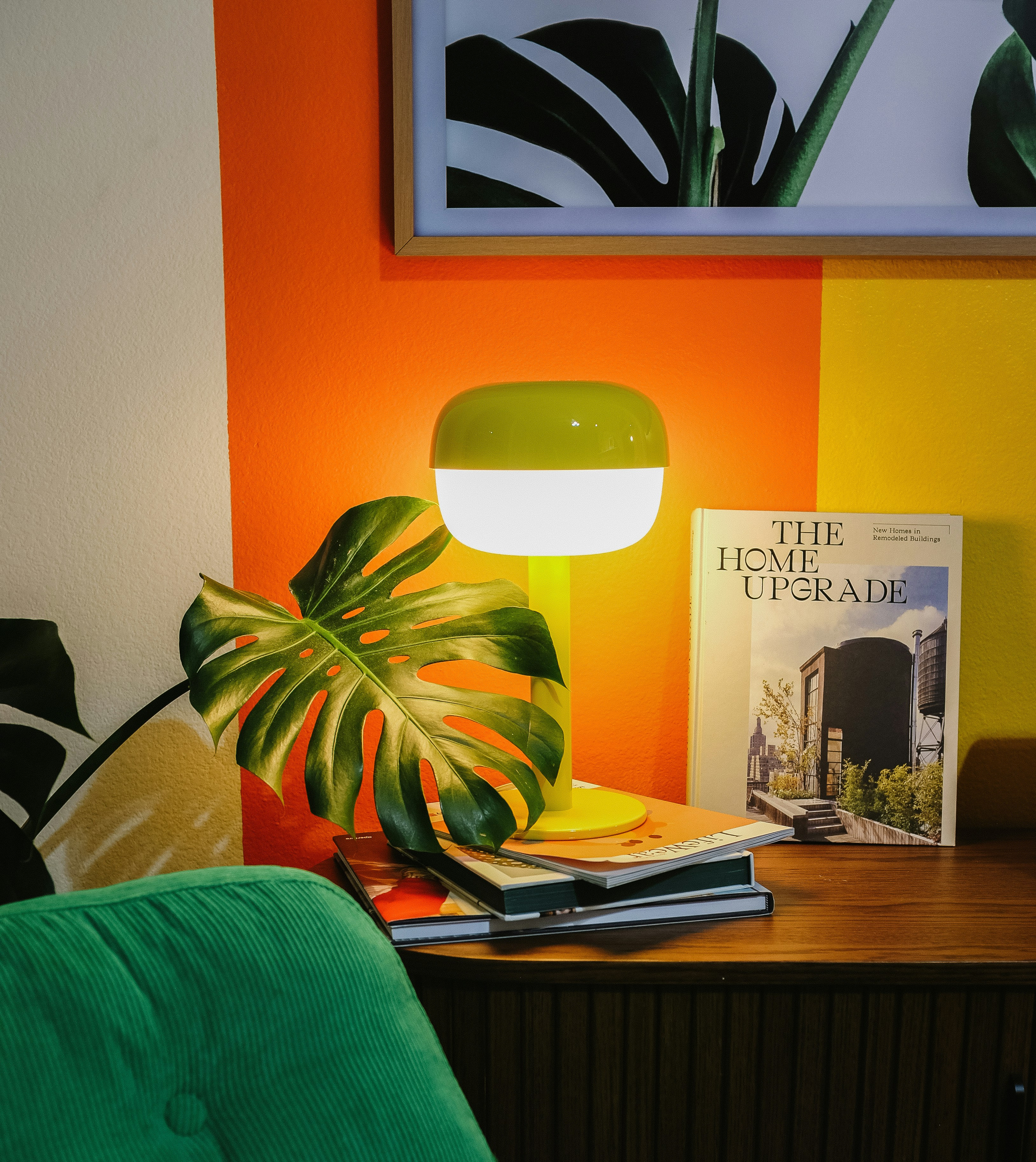 Groene fauteuil naast houten kast met plant, lamp en boek The Home Upgrade tegen oranje-gele muur.
