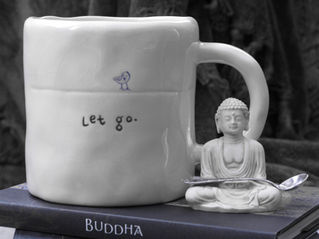 White mug with "Let go." text, bird sketch, next to a small Buddha statue holding a spoon. Mug rests on a book titled "BUDDHA". Calm mood.
