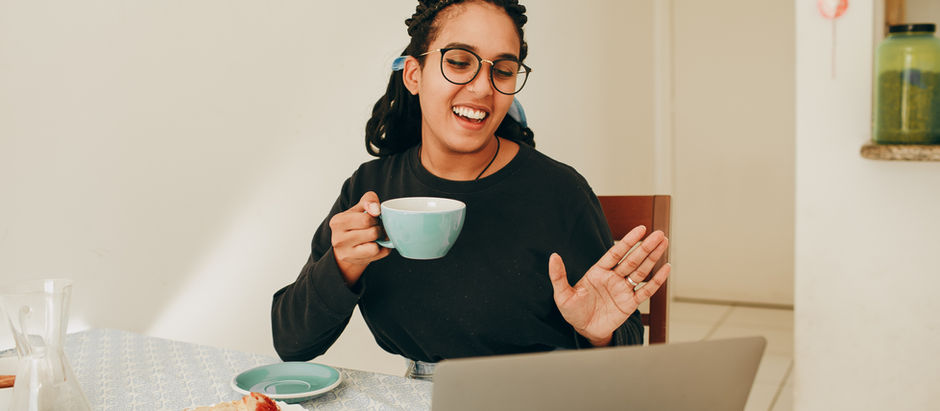 Smiling person waving at laptop, holding a turquoise cup. Bread with jam on table. Bright kitchen background, relaxed and cheerful mood.