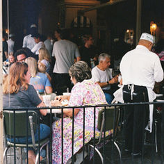 People sit and chat at outdoor café tables. A waiter in white uniform serves drinks. The café is bustling with activity and conversation.